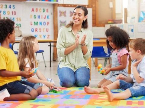 Diversity, teacher with children at school and in a classroom with a lens flare. Education or learning, happiness and smile with female person with kindergarten kids playing together in a class