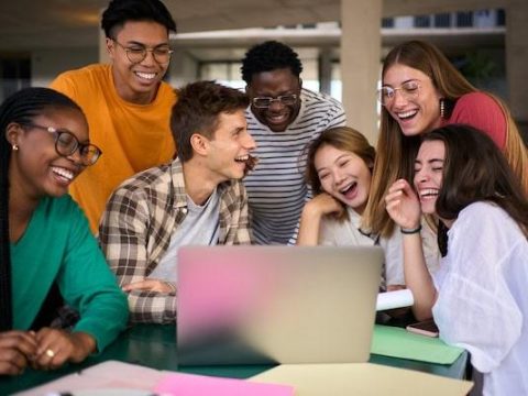 Group of young diverse students people laughing using laptop in the cafeteria on the university campus. Multiracial classmates working on a creative college project and studying together indoor