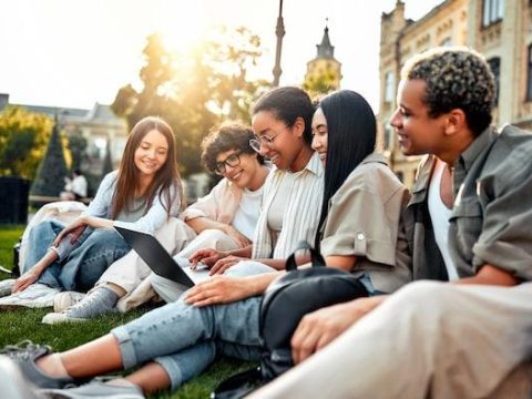 University students are sitting on the grass with a laptop and preparing for classes. Students relax on the campus.