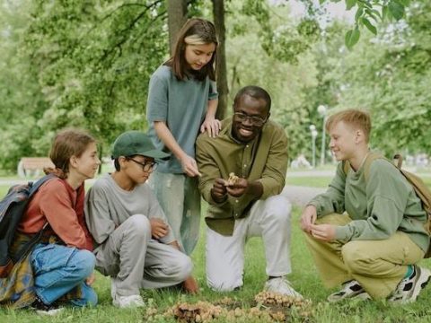 Black male young adult teacher showing mushrooms to group of diverse children and teenagers sitting and kneeling on grass in park, students observing and learning outdoors together